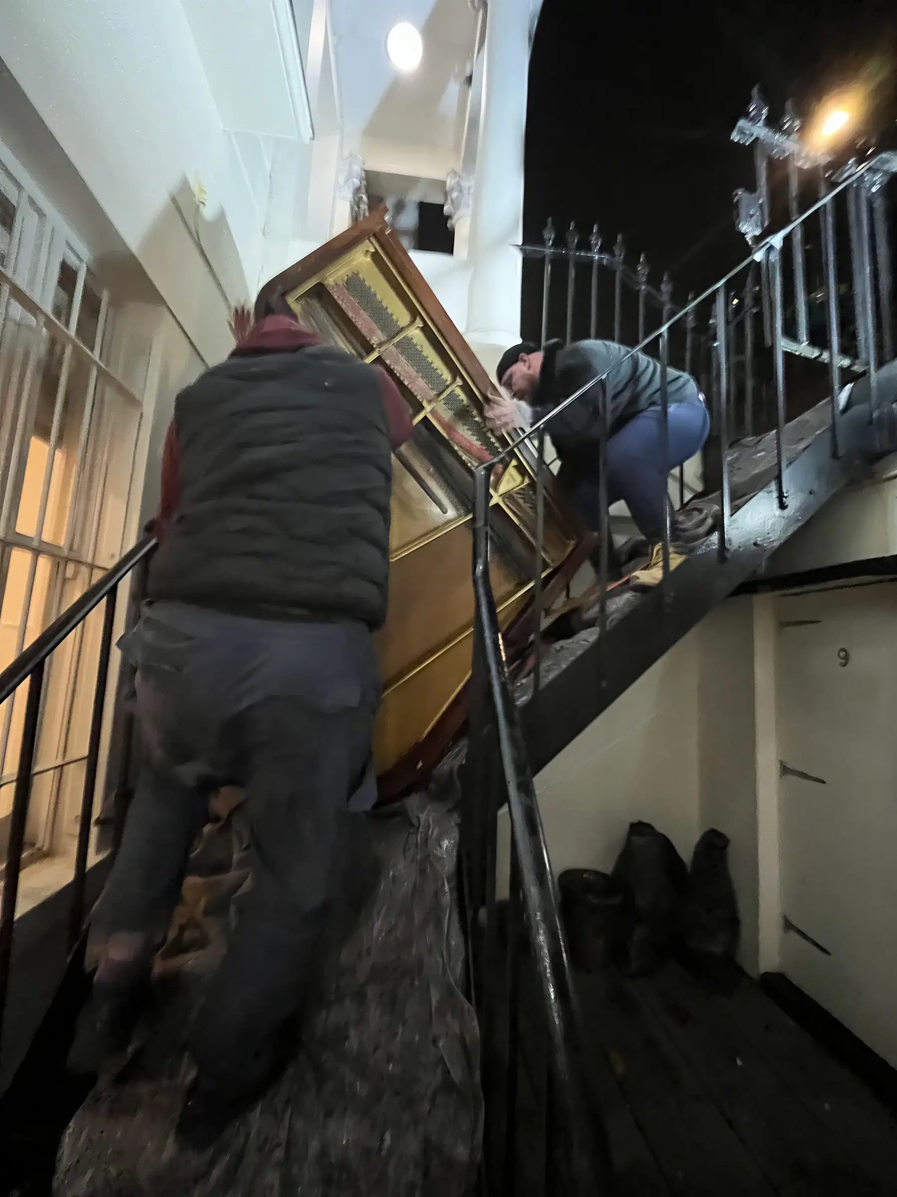 Two movers lifting a baby grand piano on its side up a narrow unlit staircase