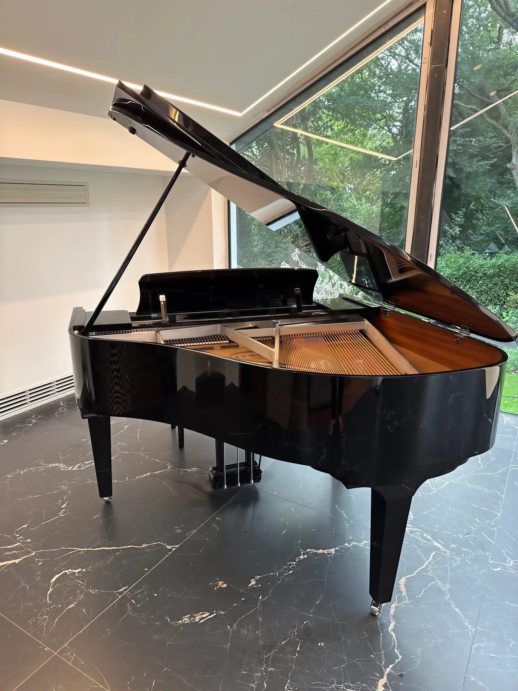 A black grand piano with the lid raised on a marble floor beside a wall of windows