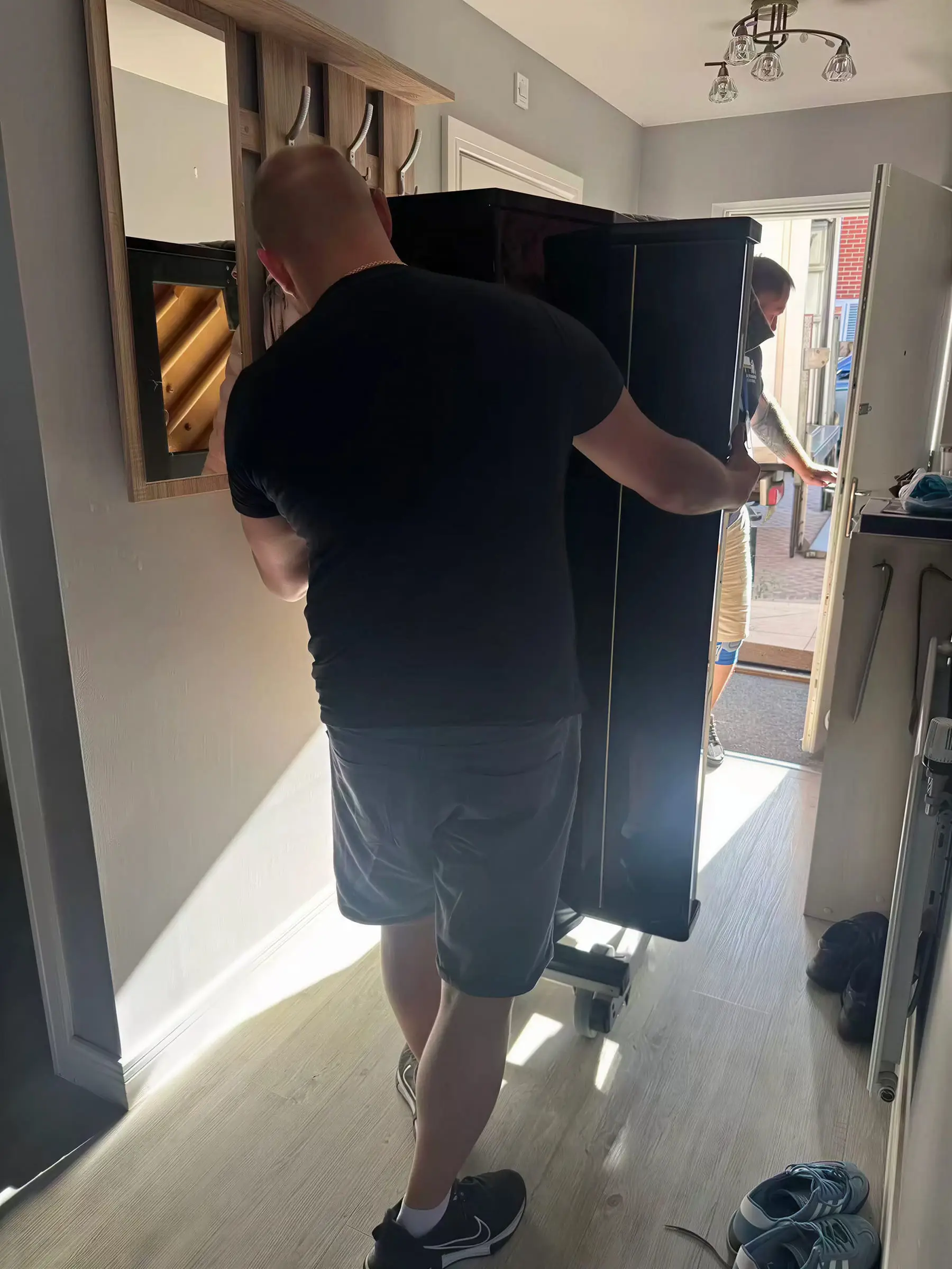A mover guiding a black upright piano through the front door of a house