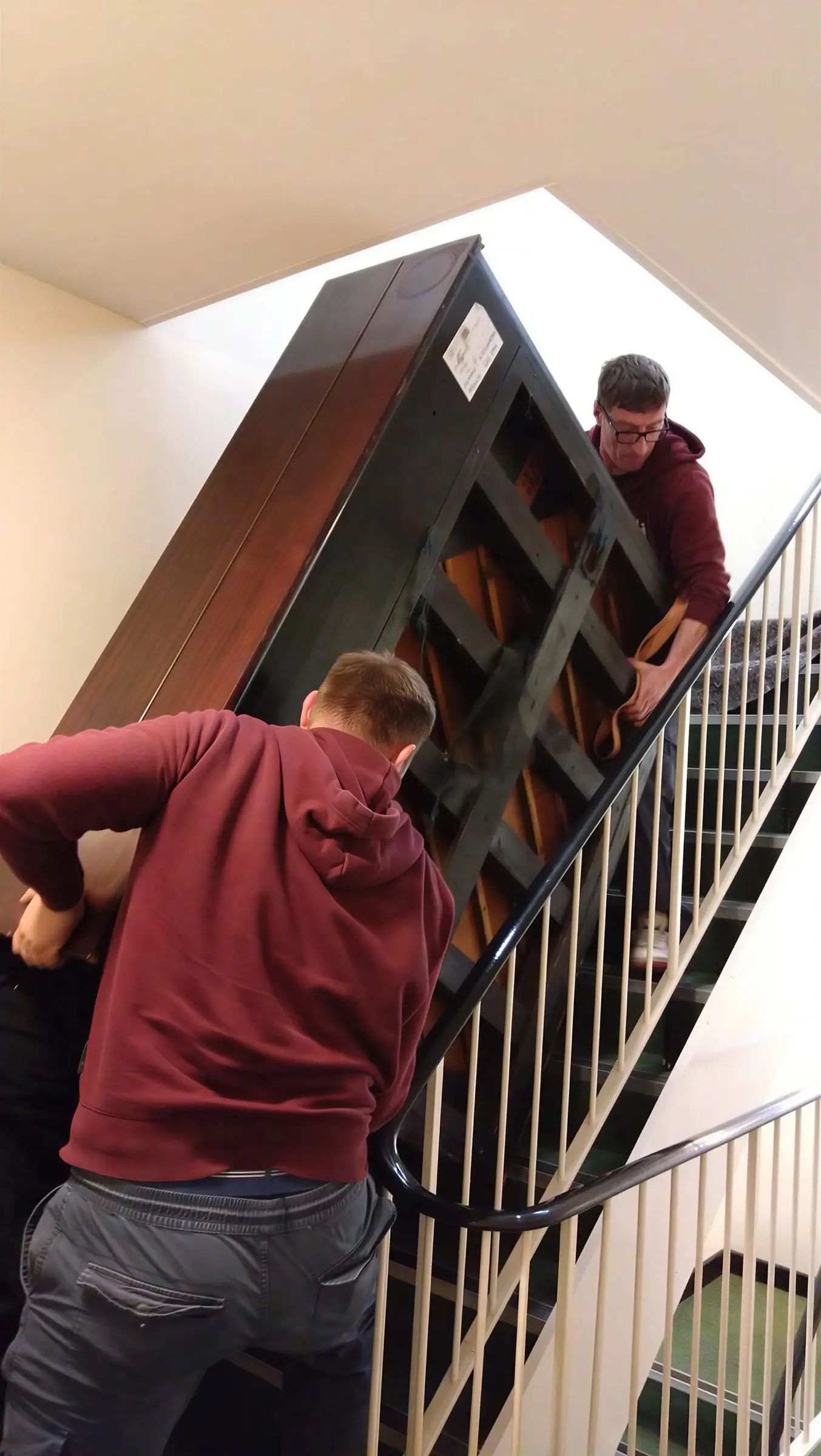 A mover carrying an upright piano up a staircase past a white banister